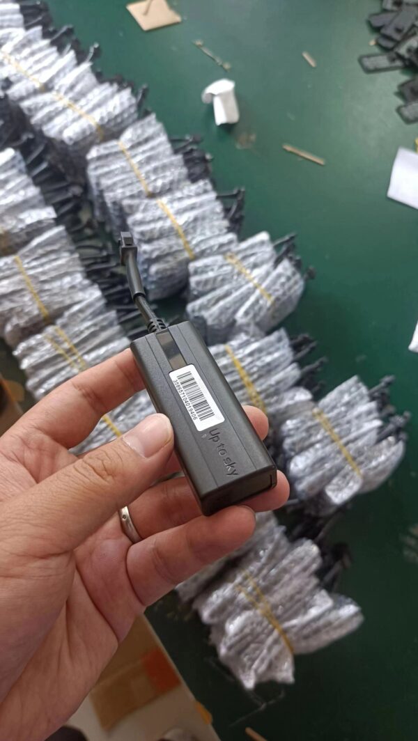 Hand holding a TK003 GPS tracker in a warehouse with rows of packaged units in the background